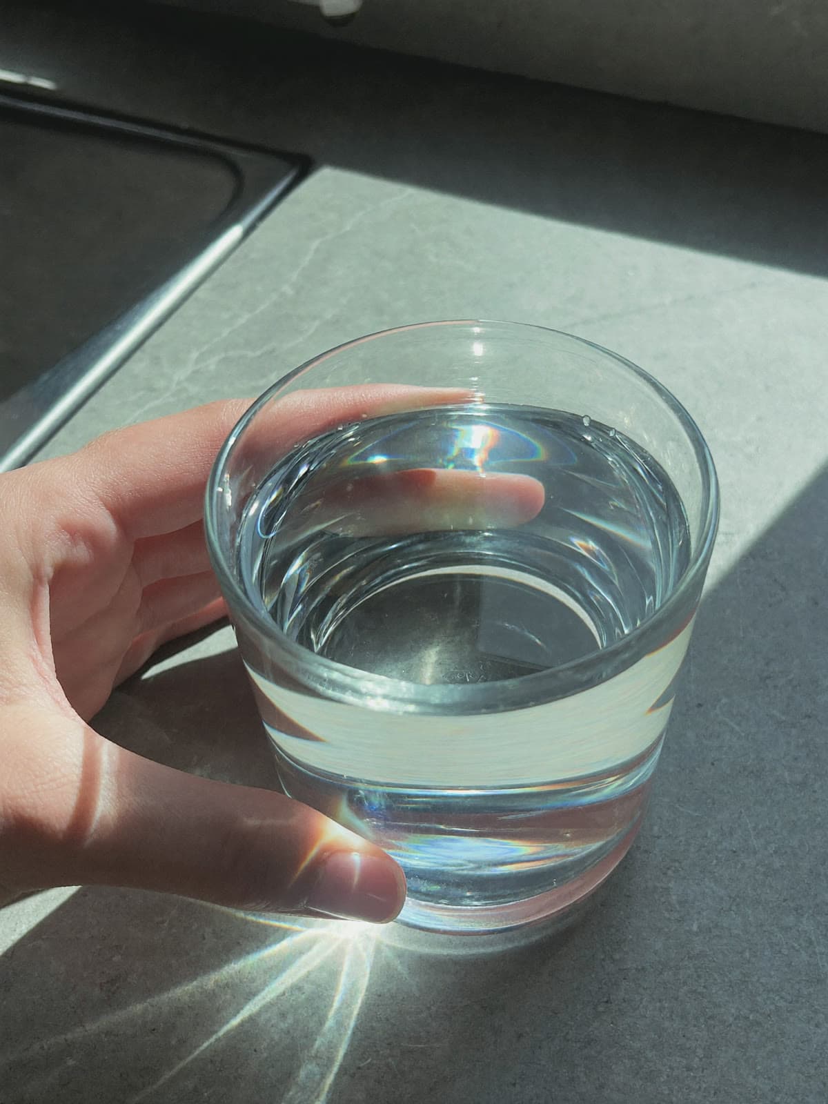 A hand lifting a glass of clear water, light refracting onto the counter